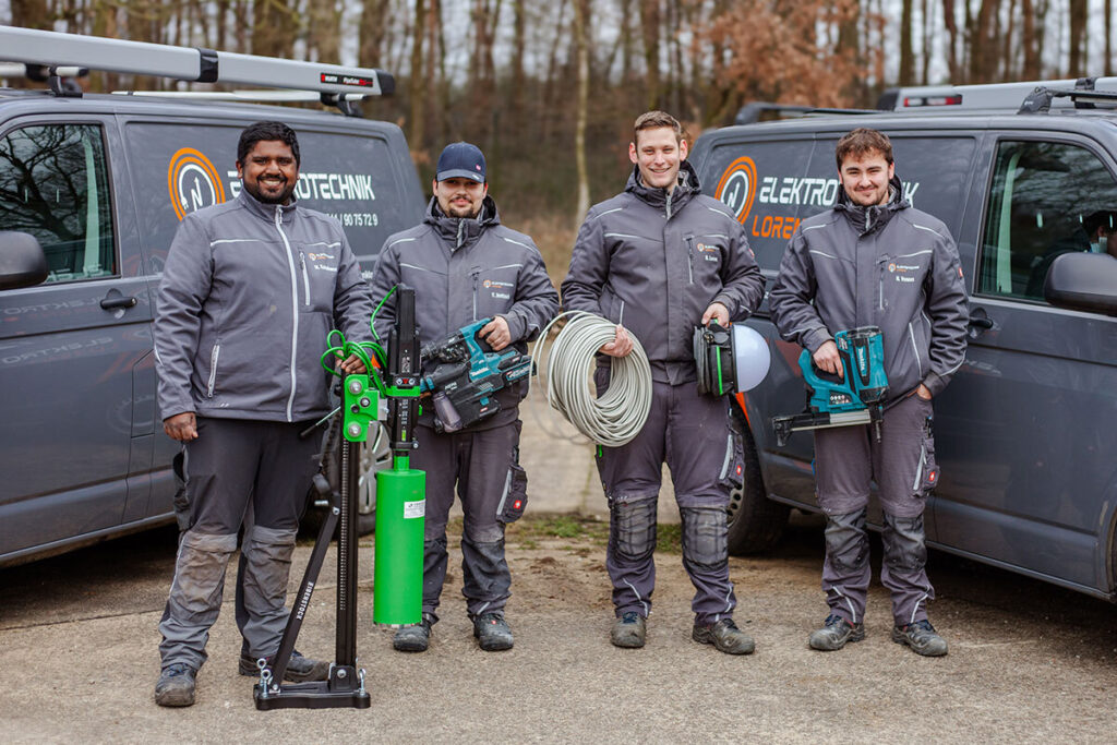 Das Team von Elektrotechnik Lorenz mit Werkzeug vor den Firmenfahrzeugen Vier Mitarbeiter von Elektrotechnik Lorenz stehen mit Werkzeug und Arbeitsmaterial vor zwei grauen VW-Transportern mit Firmenlogo